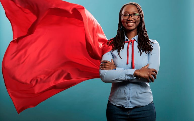 Brave and happy looking young adult superhero woman wearing red hero cape while smiling at camera. Justice defender with superpowers and mighty posture standing with arms crossed on blue