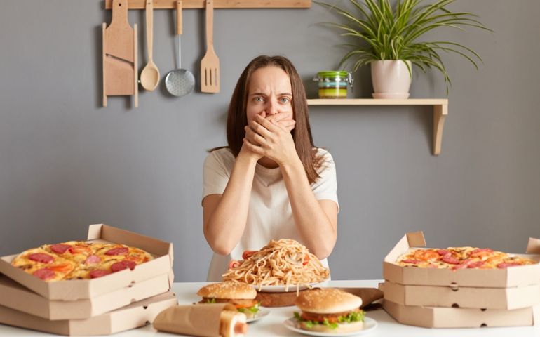 Portrait of sick Caucasian woman wearing white casual T-shirt sitting at table in kitchen, feels sick after smelling aromat of fast food, nausea, covering mouth with hands.