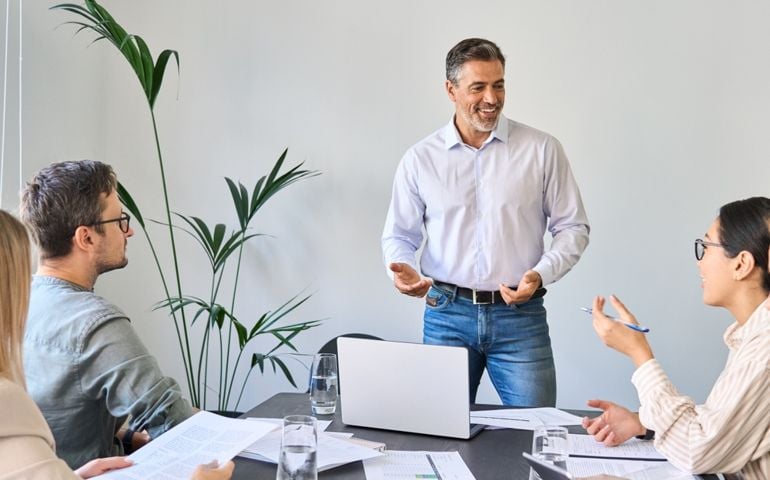 Older Latin company manager leading team meeting in office. Happy diverse professional people group working together in boardroom. Mature business man boss talking to colleagues having discussion.