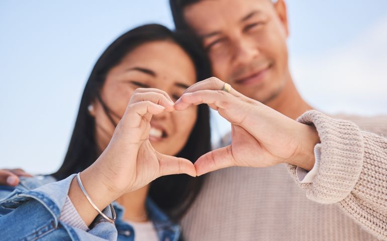 Love, heart hands and portrait of happy couple with blue sky, nature and holiday travel for summer. Care, marriage and smile, loving sign language emoji with man and woman on beach vacation together.