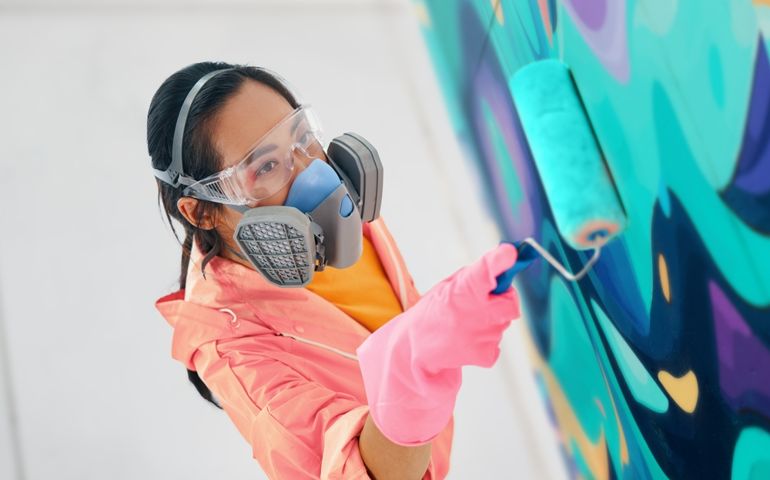 Female street artist in respirator mask painting colorful graffiti with paint roller on wall Modern art, urban concept.