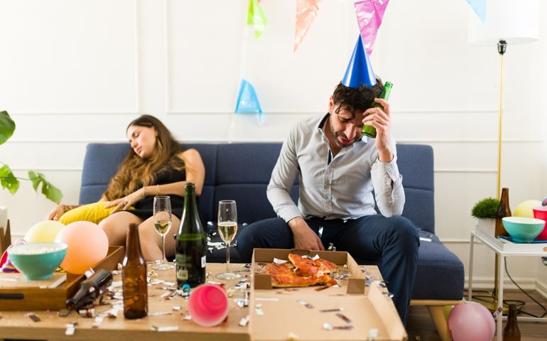 Sick man with a hangover suffering a headache while waking up to a mess and an unconscious woman sleeping on the sofa after drinking a lot of alcohol