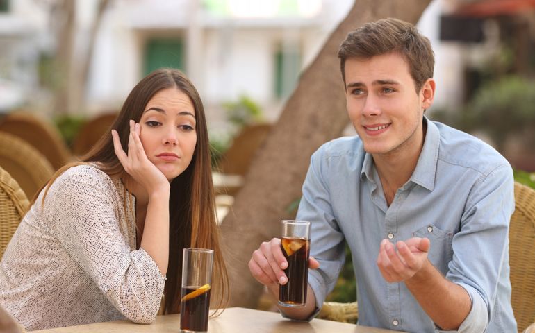 Man and woman dating in a restaurant terrace but she is boring while he telling lies with the street in the background