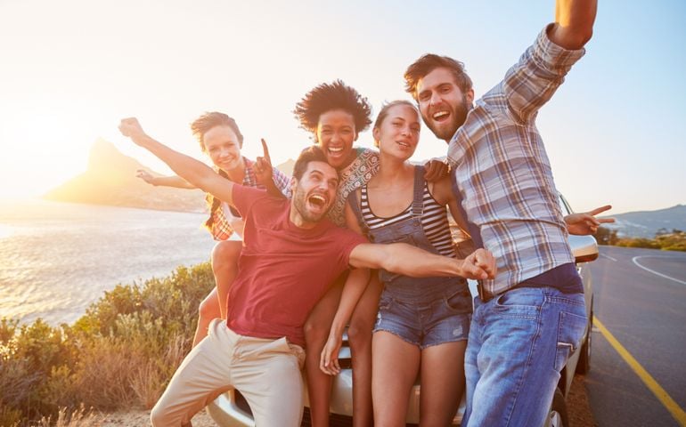 Group Of Friends Standing By Car On Coastal Road At Sunset