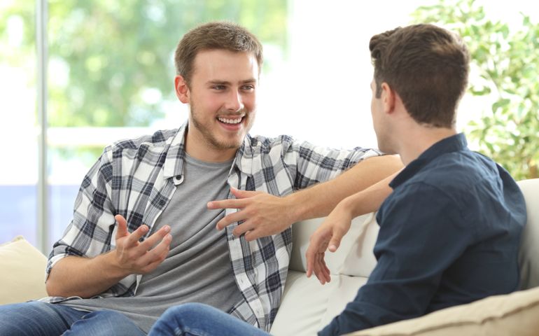 Two friends talking sitting in a couch in the living room with a window in the background at home