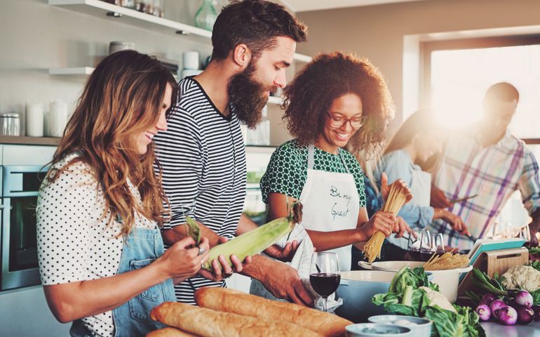 Good friends laughing and talking while preparing meals at table full of vegetables and pasta ready for cooking in kitchen