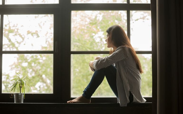 Thoughtful girl sitting on sill embracing knees looking at window, sad depressed teenager spending time alone at home, young upset pensive woman feeling lonely or frustrated thinking about problems