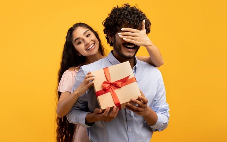 Pretty indian woman covering her boyfriend's eyes, holding wrapped gift box and greeting him with birthday or anniversary, standing over yellow studio background