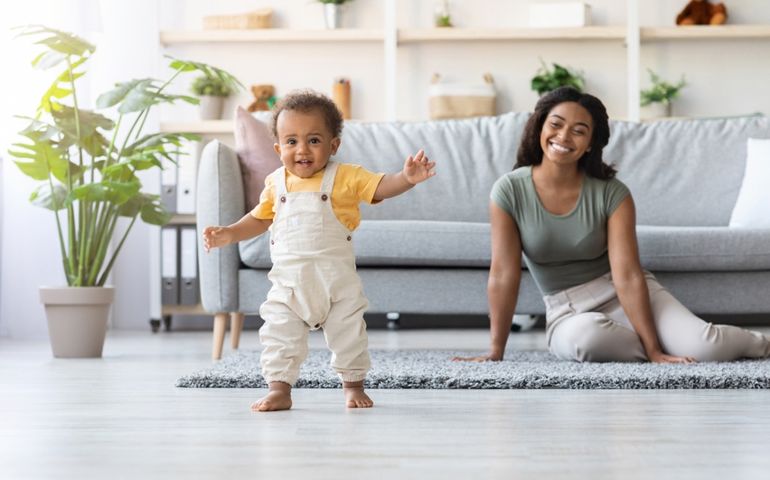 Portrait Of Adorable African American Infant Boy Making His First Steps At Home, Cute Little Black Toddler Child Wearing Jumpsuit Walking In Living Room, His Happy Mom Smiling On Background,