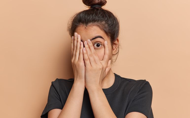 Scared Latin woman with hair bun peeks through her fingers fearfully hiding her face unable to bear sight of something terrible witnessing something truly terrible dressed in casual t shirt isolated