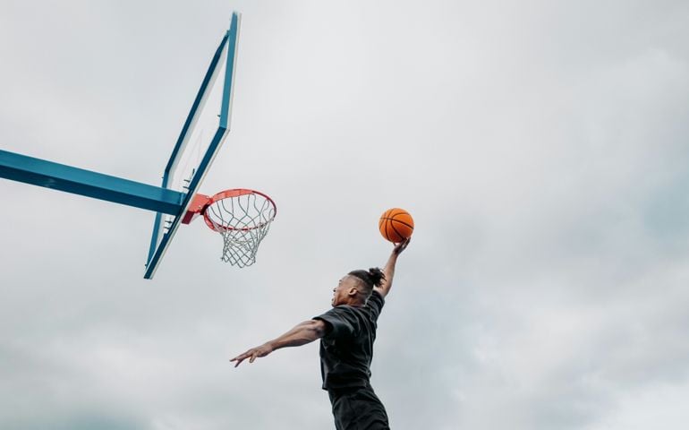 A basketball player in black athletic wear soars through the air, reaching for a powerful dunk against a backdrop of an overcast sky and a blue outdoor hoop.