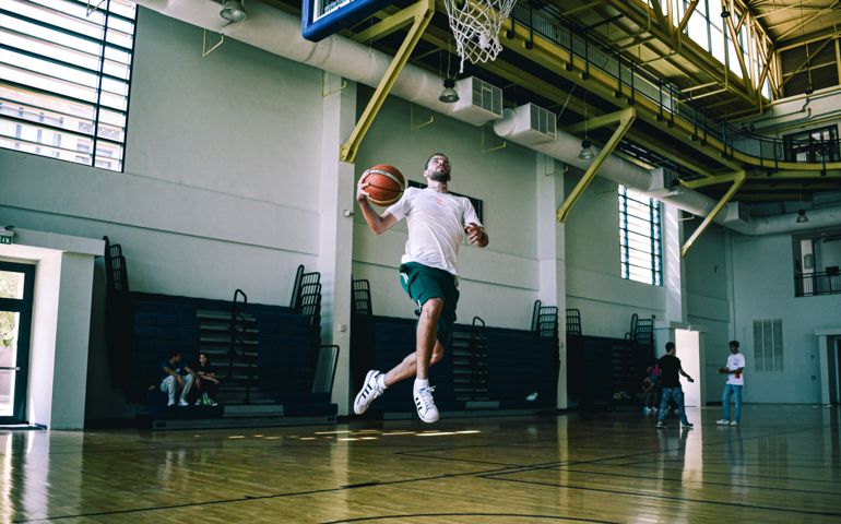 A basketball player in mid-air prepares to dunk during a game at an indoor gym, showcasing athleticism and skill on the court.