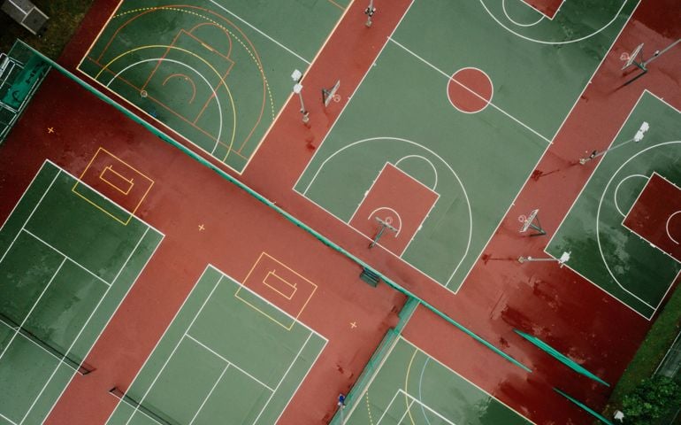 Aerial view of multiple outdoor sports courts, including basketball and tennis courts, with vibrant green and red surfaces marked by bright white and yellow lines.