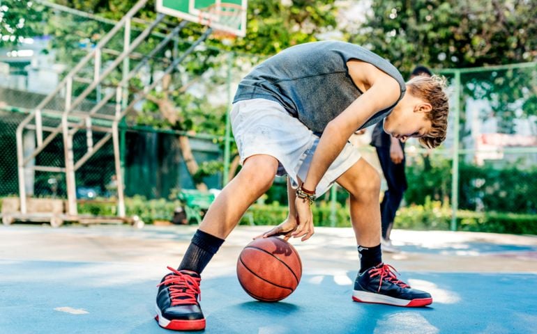 A young basketball player in a gray tank top and white shorts dribbles the ball on an outdoor court, focused on their movements while wearing black and red sneakers.