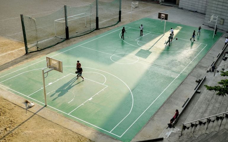 An outdoor basketball court bathed in sunlight, with players engaged in a casual game while a spectator watches from the bleachers.