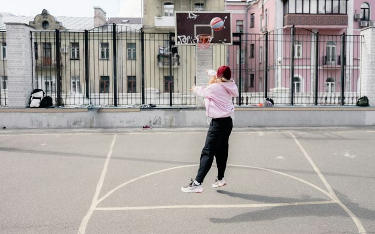 A young woman in a pink hoodie and black sweatpants takes a jump shot on an urban outdoor basketball court, with a backdrop of colorful old buildings and a graffiti-covered hoop.