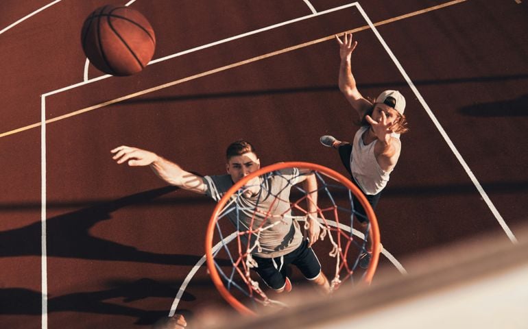 Two young men playing an intense game of basketball on an outdoor court, captured from a dramatic overhead perspective as one player attempts a layup while the other tries to block the shot.