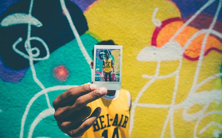 A person wearing a yellow ‘Bel-Air Academy’ basketball jersey holds up a Polaroid photo of themselves in front of a colorful graffiti wall, creating a layered visual effect.
