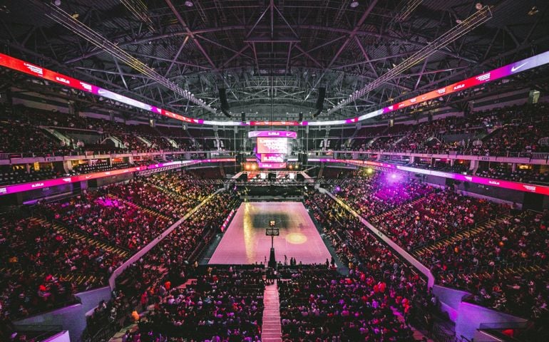A packed indoor basketball arena illuminated with vibrant lights as fans fill the stands, creating an electric atmosphere for the game.