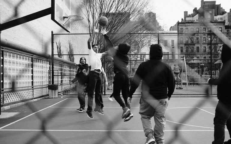 A black-and-white photo captures an intense basketball game on an urban outdoor court, framed by a chain-link fence, with players leaping for the ball against a city backdrop.