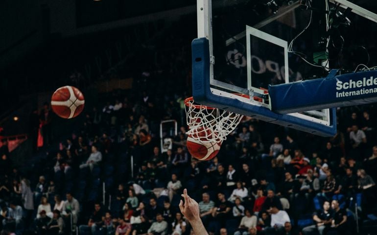 A basketball soars through the air and swishes into the net during warm-ups before a professional game, with a packed arena of spectators watching in the background.