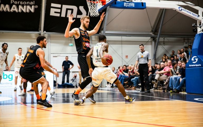 Duane Wilson of Nürnberg Falcons (white) drives past Marko Bacak of wiha Panthers Schwenningen (black) during a BARMER 2. Basketball Bundesliga ProA game, with a crowd of spectators watching the action.