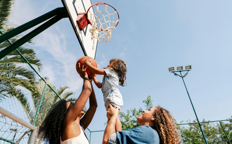 A young child in a light blue outfit is lifted by two women to shoot a basketball into the hoop on an outdoor court, surrounded by palm trees and a clear blue sky
