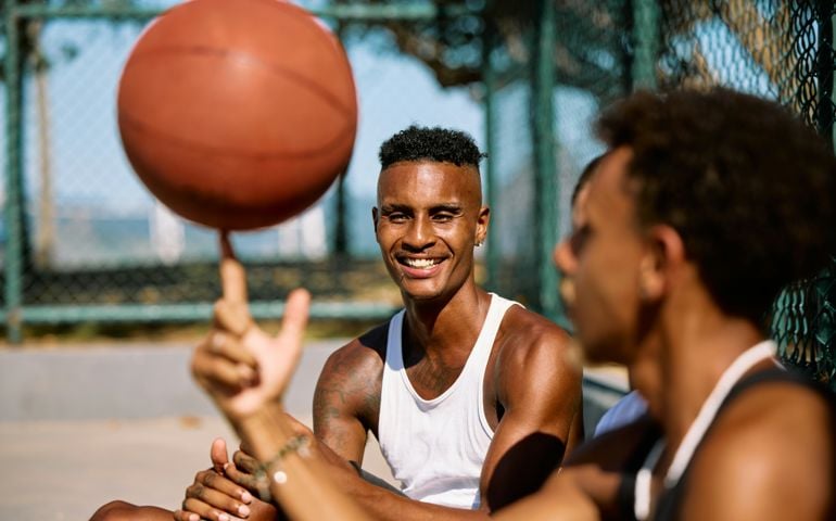 Two young men enjoy a casual basketball game on an outdoor court, with one spinning a basketball on his finger while the other smiles in the sunlight.
