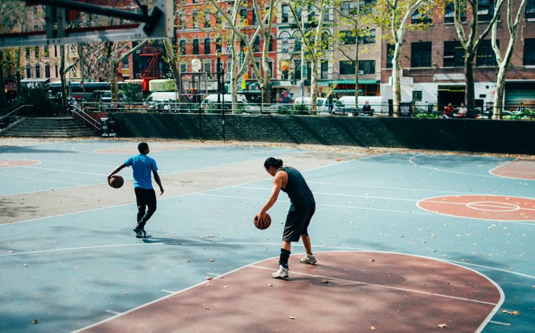 Two people playing basketball on an outdoor court in an urban setting, surrounded by trees and city buildings, with fallen leaves scattered across the blue and red playing surface.