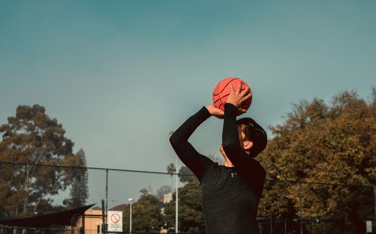 A basketball player in a black long-sleeve shirt prepares to take a shot on an outdoor court, framed against a clear sky and autumn-colored trees in the background.