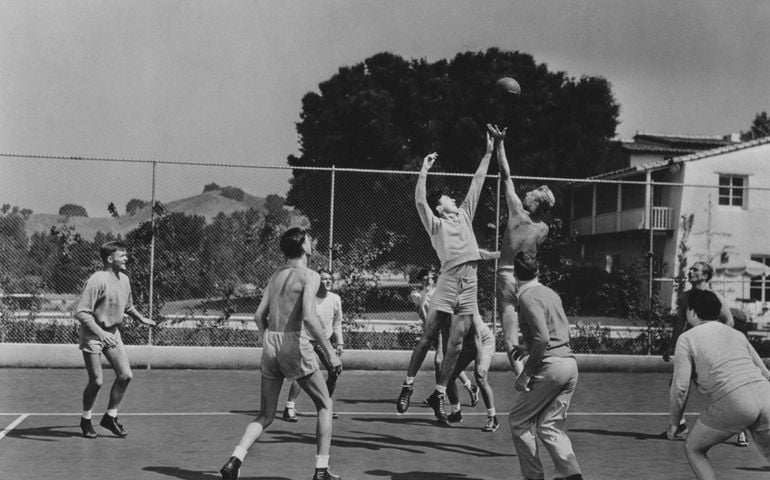 A vintage black-and-white photograph of an outdoor basketball game captures players in mid-action, leaping for a jump ball, with a backdrop of hills, trees, and a residential area.
