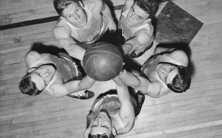 Black and white overhead shot of five vintage basketball players reaching for the ball during a game.