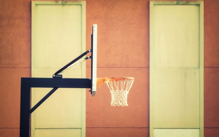 A basketball hoop and backboard stand against a warm-toned wall, framed by two large, muted green windows, creating a minimalist and nostalgic urban court setting.