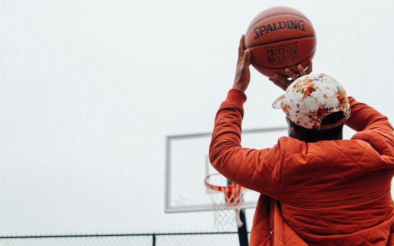 A basketball player in a red quilted jacket and floral cap prepares to take a shot on an outdoor court, focusing on the hoop in the distance.