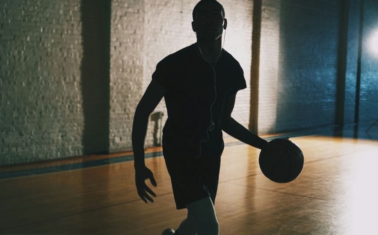 A silhouette of a basketball player dribbling on an indoor court, illuminated by dramatic lighting that highlights the intensity and focus of the game.