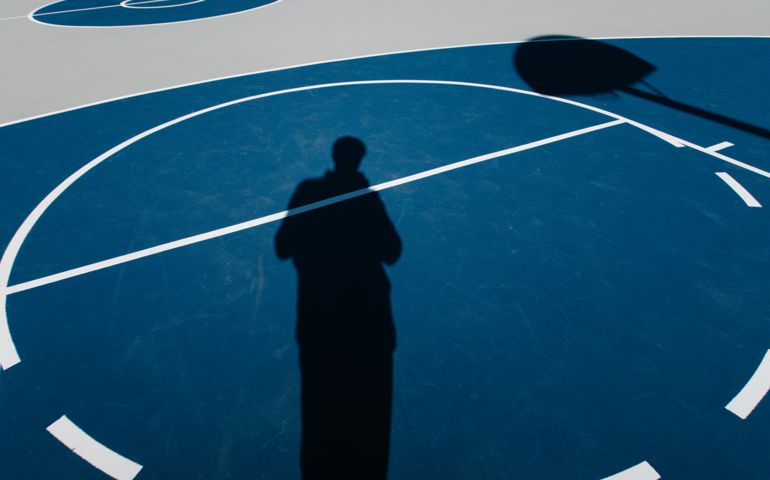 Shadow of a person on a blue outdoor basketball court, with the hoop’s silhouette cast onto the playing surface, creating a striking minimalist composition.