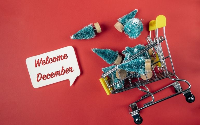 A miniature shopping cart tipped on its side spills tiny Christmas trees onto a red background, next to a speech bubble sign that reads "Welcome December"—hinting at the festive surprises and hidden history packed into the last month of the year.