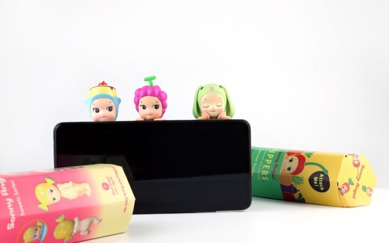 Three Sonny Angel collectible figures with whimsical costumes—grape, frog, and birthday cake—perch atop a phone, with brightly colored packaging scattered nearby; a playful, surreal vibe echoes the quiz’s theme of finding your matching Sonny Angel energy