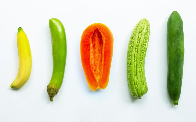 A row of five fruits and vegetables is arranged upright on a white background: a banana, green eggplant, halved papaya, bitter melon, and cucumber. The central placement and shapes suggest visual innuendo without showing anything explicit.
