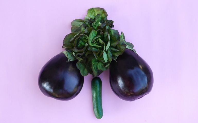 Two eggplants and a head of lettuce are arranged to resemble the male anatomy, with a small cucumber positioned below on a pastel pink background. The playful, suggestive layout creates a cheeky visual without being explicit.