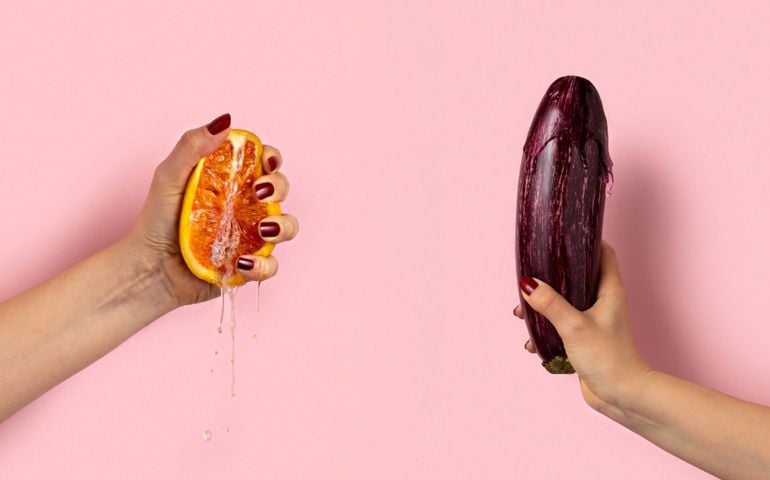 Two hands hold suggestive fruits against a pink background — one squeezes a juicy half of a grapefruit, with liquid dripping down, while the other grips a striped eggplant upright. Both hands have burgundy nail polish, enhancing the playful, innuendo-driven composition.