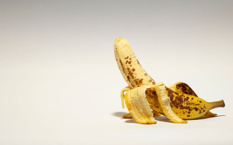 A ripe, speckled banana is partially peeled and propped upright on a neutral background. Its positioning and exposed shape create a humorous, suggestive visual commonly associated with innuendo.