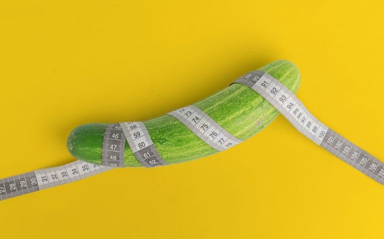 A long, curved cucumber is wrapped with a measuring tape against a bright yellow background. The playful composition and suggestive placement hint at innuendo without showing anything explicit.