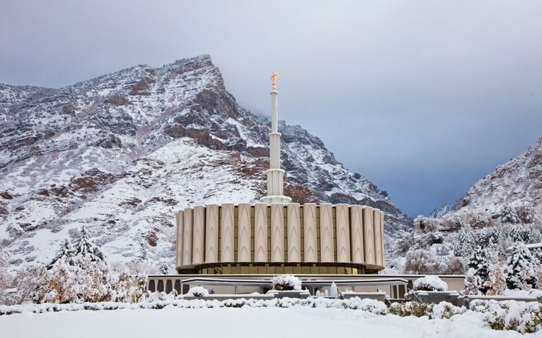 A snow-covered LDS temple with a modern circular design stands in front of a rugged mountain backdrop. The temple’s spire is topped with a golden angel Moroni statue, and surrounding trees are dusted with fresh snow under a cloudy sky.